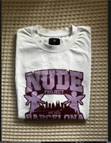 Remera blanca con estampado gráfico en colores morado y rosa que incluye el texto 'NUDE PROJECT' y 'BARCELONA'.