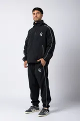 Pantalón de jogging negro de taslan con corte holgado, cintura elástica con cordón ajustable, bolsillos laterales y puños elásticos. Presenta una franja blanca a lo largo de la pierna y logo bordado en blanco.