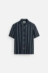 Camisa azul marino con rayas verticales blancas, corte relaxed fit, cuello bowling, manga corta, bajo con aberturas laterales y cierre frontal con botones.