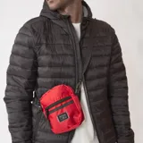 Morral pequeño tipo bandolera, color rojo brillante, con textura tejida y dos cierres frontales negros. Incluye un parche rectangular con texto en la parte inferior.