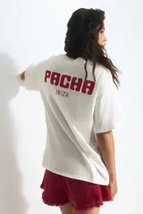 Remera blanca de algodón de corte amplio con cuello redondo y mangas cortas. Estampado frontal de dos cerezas rojas con hojas verdes y el logo de la marca Pacha en rojo.