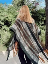 Ruana tejida a mano en telar, confeccionada en lana merino con diseño a rayas horizontales en tonos blanco, marrón, verde y negro. Presenta flecos en los bordes.