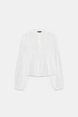 Camisa blanca de escote pico y manga larga con puños elásticos. Detalle de puntilla combinada a tono y cierre frontal con botones.