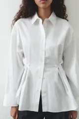 Camisa blanca de cuello solapa y manga larga. Detalle de pliegues con botones. Cierre frontal con botones.