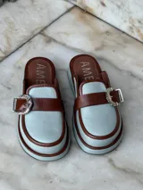 Zuecos negros de cuero con suela gruesa, detalle de tira de gamuza con hebilla metálica plateada y logo "AMÉN" estampado en la plantilla.