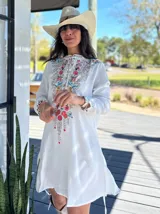 Vestido corto blanco con bordado floral multicolor en el frente, escote redondo con cierre de cremallera parcial y mangas largas.