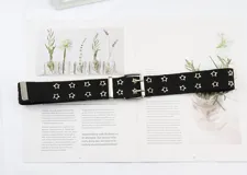 Cinturón de lona negro con doble hilera de tachas metálicas en forma de estrella de cuatro puntas. Cuenta con hebilla doble de metal plateado y pasador metálico.