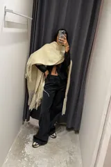 Ruana o poncho de tejido grueso color beige claro, con flecos largos en el borde inferior y ribete de costura en contraste en la abertura frontal.