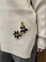 Sweater de punto color beige con cuello alto, mangas largas y corte holgado. Presenta tajos laterales y bordados florales en puños y cadera.