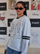 Buzo blanco de manga larga con cuello redondo y puños y ruedo en rib negro. Presenta un gran estampado frontal de la letra 'M' rellena de dibujos de personajes de Disney, incluyendo a Mickey Mouse. Las mangas tienen dos franjas negras a la altura del bíceps.