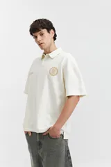 Remera polo color beige con textura acanalada, corte oversize, cuello clásico con botones y logo estampado en el pecho.