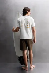 Camiseta blanca de corte holgado confeccionada en tejido de algodón con elasticidad. Tiene cuello redondo, manga corta y estampado en el frente con el logo de la marca y un diseño de tablas de surf.