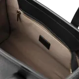 Cartera negra de simil cuero graneado con doble asa de mano y correa bandolera regulable. Tiene cierre superior, dos bolsillos internos abiertos y uno con cierre. El interior es de tela de algodón y poliéster.