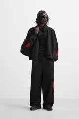 Chaqueta negra con detalles en rojo, cuello alto, cierre frontal con cremallera, bolsillos laterales y puños elásticos.