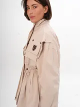 Trench color beige con cuello alto, cierre con botones a presión, bolsillos con solapa y cordón ajustable en la cintura.