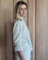 Blusa color beige con cuello con volados, mangas largas abullonadas con puños elásticos y cierre con botones en la parte delantera.