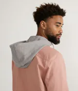 Camisa overshirt color salmón de sarga de algodón con capucha gris ajustable con cordones. Tiene cierre frontal con botones, dos bolsillos con solapa en el pecho y mangas largas con puños abotonados.