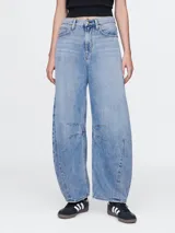 Jean de tiro alto con corte horseshoe, caracterizado por su silueta curva y amplia en las piernas. Presenta costuras reforzadas en la zona de las rodillas y un lavado en tono azul medio.