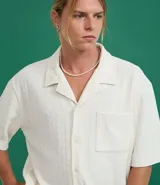 Camisa de jacquard color crema, de corte oversized, con cuello solapa, manga corta, abotonadura frontal, bolsillo en el pecho y textura en relieve.