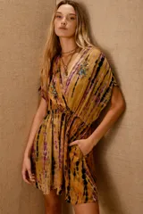 Vestido corto con estampado tie-dye en tonos mostaza, morado y negro. Presenta escote cruzado en V, mangas cortas tipo kimono y cintura ajustada con elástico. Tiene pequeños bordados de cuentas de colores en el escote.