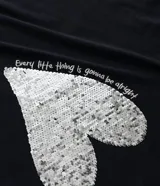 Remera negra de algodón con cuello redondo y manga corta. Presenta un estampado frontal de un corazón dorado de lentejuelas reversibles y texto blanco.