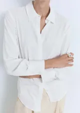 Camisa blanca de tejido fluido, corte recto, cuello camisero, cierre frontal con botones y mangas largas con puños abotonados.