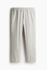 Pantalón de vestir gris claro, de corte slim fit, con cintura elástica y cordón ajustable. Presenta bolsillos laterales y traseros ribeteados, pliegues cosidos y doblez en el bajo.