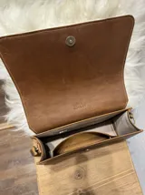 Bandolera de cuero color negro con forro interior y cierre. Presenta un cuerpo cubierto de lana de oveja color blanco y correa larga de cuero marrón con hebilla ajustable.