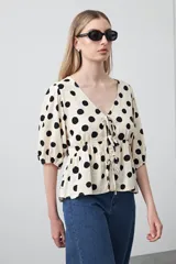 Blusa de silueta relajada con estampado de lunares negros sobre fondo crema. Cuenta con escote en V con lazos regulables, frunce bajo el busto y mangas cortas con volumen.