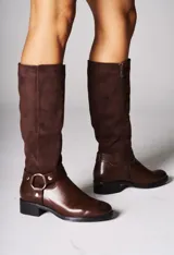 Bota de caña alta color marrón, con combinación de cuero liso en la parte inferior y gamuza en la caña. Tiene un detalle de arnés con hebilla dorada a la altura del tobillo y cierre lateral.