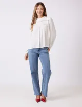 Blusa blanca de gasa plisada con encaje calado en el cuello y el pecho. Cuello a la base fruncido, manga larga con puño elastizado y espalda lisa con abertura tipo gota y botón.