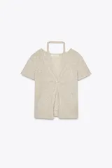 Camiseta color beige de tejido de punto, con cuello halter y escote pico. Tiene cierre frontal con botones y mangas cortas.