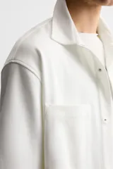 Sobrecamisa color beige de tejido elástico, con cuello solapa, manga corta y bolsillos de parche en el pecho. Cierre frontal con botones a presión.
