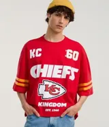 Remera roja de algodón con estampado del logo de los Kansas City Chiefs, el nombre del equipo y el número 60 en blanco. Tiene mangas cortas con rayas amarillas y rojas.