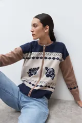 Chaqueta de tejido punto con cierre frontal, cuello redondo y manga larga. Presenta un diseño jacquard en tres colores: azul marino, beige y marrón camel. El cuerpo principal es beige con motivos de rosas azules y patrones geométricos en azul marino. Los hombros y mangas son de color marrón camel con patrones geométricos en azul marino. El cuello y el elástico inferior son de color azul marino.