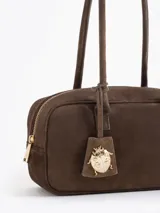 Cartera rectangular de estructura firme confeccionada en cuero descarne vacuno color marrón. Presenta manijas fijas, cierre superior y un charm metálico dorado en forma de vaquita de San Antonio colgando de un tag lateral. Incluye el logo de la marca grabado en relieve en el frente.