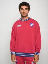 Buzo de felpa color rojo con efecto lavado, cuello redondo con ribete azul y blanco, puños y cintura acanalados con ribete azul y blanco, escudo del Club Nacional de Football bordado en el pecho y logo de Umbro bordado en la manga.