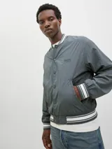 Campera bomber casual color gris verdoso, con cuello tipo baseball y puños de canalé a rayas blancas. Cierre con botones a presión y bolsillos frontales. Confeccionada en tejido liviano y transpirable con hombros caídos.