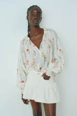 Blusa oversized de viscosa color crema con estampado floral. Tiene cuello corto con volado y abertura en pico con tiras finas. Las mangas son raglán largas con elástico fino en los puños y el bajo es redondeado.