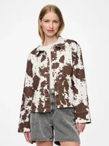 Chaqueta de jean con estampado animal print de vaca en tonos marrones y blancos, corte recto, cuello camisero y cierre frontal con botones.