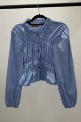 Camisa celeste a rayas verticales blancas, de corte entallado, con cuello clásico, cierre frontal con botones rojos, frunces en el delantero y mangas largas abullonadas con puños elásticos.