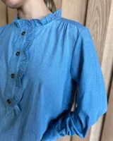 Camisa a rayas verticales color beige y blanco, de corte holgado, con cuello con volados, cierre parcial con botones y mangas largas con puños abotonados.