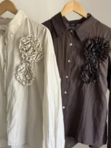 Camisa de corte clásico, color marrón oscuro con brillo sutil dorado, confeccionada en tela nacarada. Presenta apliques de flores tridimensionales fruncidas en el área del pecho.