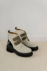 Botines de cuero color crema con puntera redondeada y suela track negra. Presentan dos tiras horizontales de cuero verde oliva con cierre de velcro.