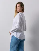 Camisa blanca de lino y rayón, con cuello a la base y escote en V abotonado. Tiene dos bolsillos con botón en el pecho y mangas largas regulables con presilla y botón.