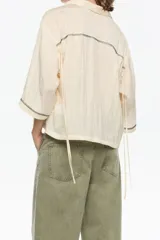 Camisa corta de corte boxy, confeccionada en tejido fantasía texturado color crema o amarillo muy pálido. Presenta cuello camisero, botones frontales y manga 3/4 con sisa ajustable mediante tiras de tejido, además de pespuntes decorativos negros que realzan el borde de las mangas.