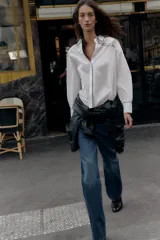 Camisa blanca de popelín con vivos negros a contraste en el cuello, el frente y los puños. Presenta un corte holgado y mangas largas con puños anchos.