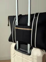 Bolso tipo tote de neopreno color negro y marrón, con cierre superior, bolsillo frontal marrón con logo rojo, bolsillo interno y externo con cierre, doble asa de mano marrón con detalles blancos y opción de correa larga.