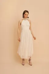 Vestido midi de fiesta color crudo, con cuello halter, cuerpo drapeado y falda de microtul plisado.