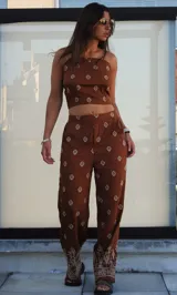 Conjunto de dos piezas color marrón chocolate, compuesto por un top corto de tirantes finos y un pantalón ancho de tiro alto. Ambas prendas presentan un estampado geométrico en color blanco o beige claro, con un diseño de borde más elaborado en el bajo del pantalón.